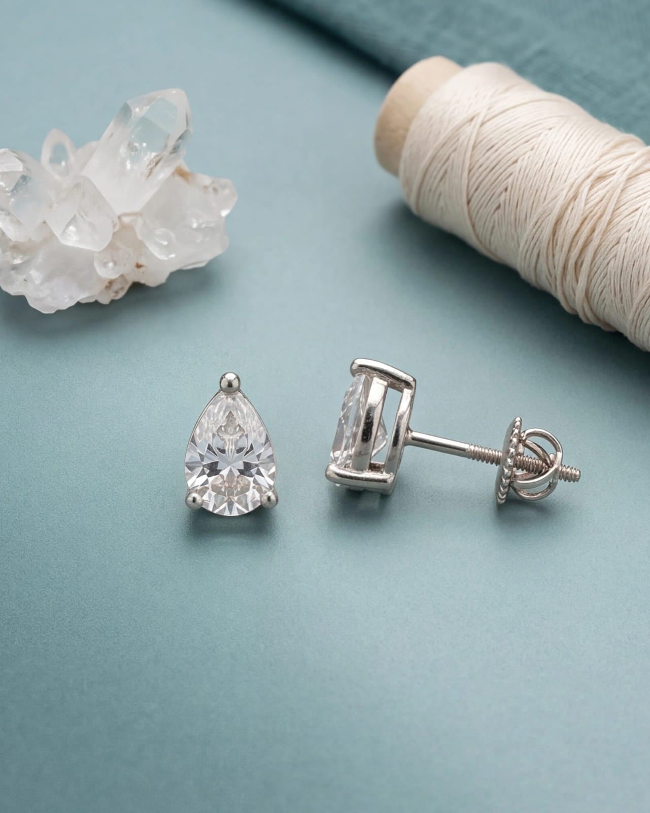 Pair of silver teardrop earrings on a light blue surface with a crystal and spool of thread. #color_silver